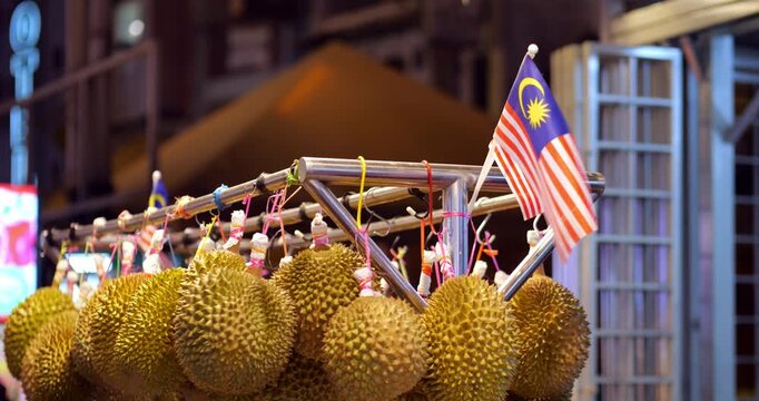 Ripe durian fruits hanging for sale at a night market stall in Malaysia, with small national flag waving, showcasing a popular and iconic Southeast Asian street food scene