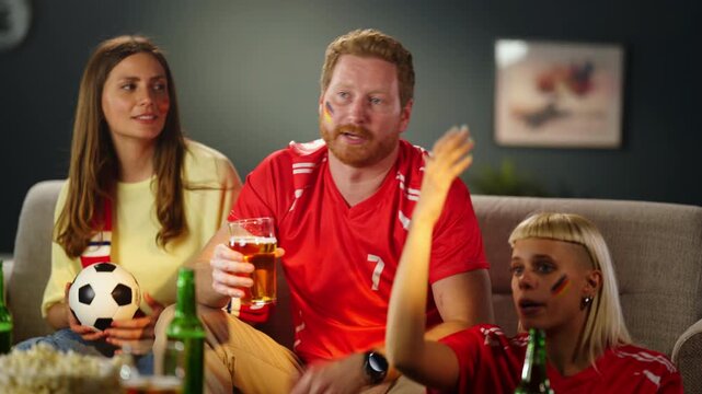 Group of friends gather in a living room to watch a soccer match. They cheer, drink beverages, and show support for their favorite team. The atmosphere is lively and energetic.