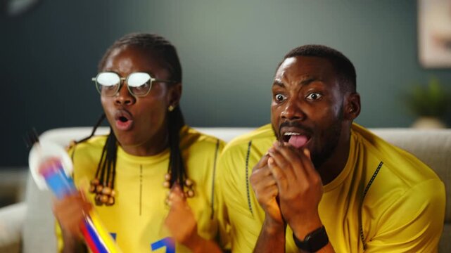 Two fans watch a sports game together at home. They cheer and react to the happening moments. Their matching shirts show their team spirit and support.