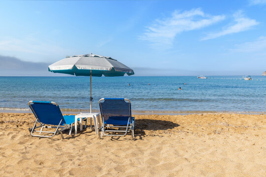 Relaxing summer sand beach with blue sun loungers and silver umbrella by calm sea after low tide on Corfu island, Greece.