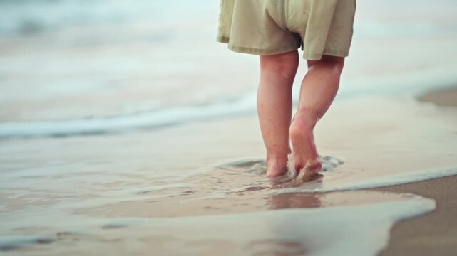 Little Asian toddler girl walking barefoot on a sandy beach watching sea waves during a summer family vacation. Close-up of child's feet on the shore. Concept of childhood discovery, nature