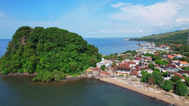 Aerial drone footage of Pecaron village nearby the white sand beach, with Maulana Ishaq mosque, a little peninsula hill with forests, in Situbondo regency, Java island, Indonesia, blue sea water
