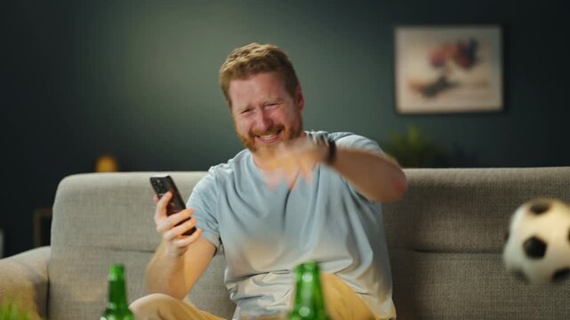 Excited football supporter checking betting app on smartphone while watching live sports match at home with soccer ball and drinks nearby.