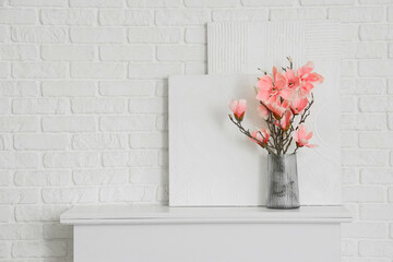 Naklejka premium Vase with artificial magnolia flowers and paintings on fireplace near white brick wall