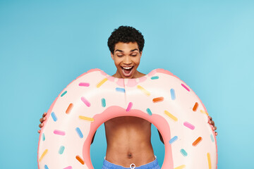 Naklejka premium Joyful young man holds a giant donut float against a vibrant blue backdrop at summer pool party