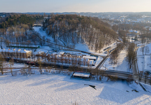 Aerial view of a snow-covered landscape featuring a frozen river, a road bridge, and a wooded hill under bright winter sunlight in Vilnius County, Lithuania.