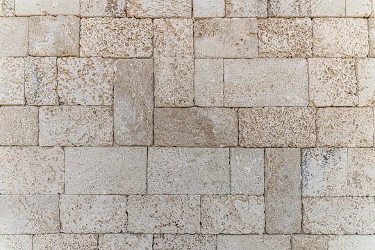 Close-up of a natural limestone brick wall with rough porous stone blocks in neutral beige tones. Architectural stone texture background with geometric pattern and rustic Mediterranean style.