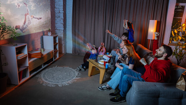 Happy family watching soccer projection at home during football fan night. Concept of live sports streaming, home cinema, match day party, family entertainment, and subscription media ads.