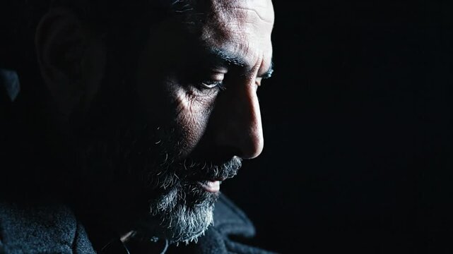 Low key shot of a pensive man with a grey beard looking downward in thought against a dark background.