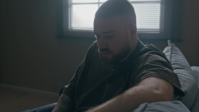 Tilt up shot of Caucasian bearded man looking through overdue bills while sitting with laptop on couch in apartment, appearing confused and stressed
