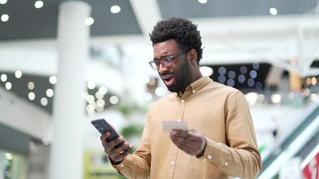 Shocked upset african american man checks his credit card on mobile phone standing in mall shopping center. Frustrated male faced fraud or glitch and error with banking. He became victim of deception