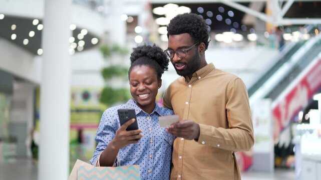 Happy african american family couple shoppers using mobile phone and credit card to make online payments standing in shopping center mall store. A man and woman uses smartphone doing purchase paid app