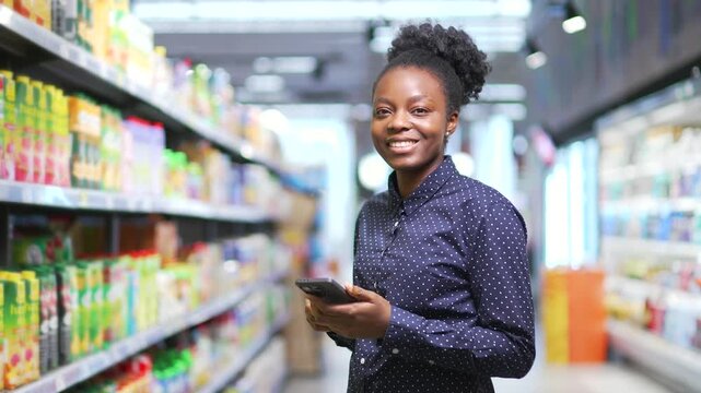 Portrait of young smiling African American woman in grocery store in supermarket. A happy female shopper uses a mobile phone standing between rows of shelves in market, shopping and looking at camera