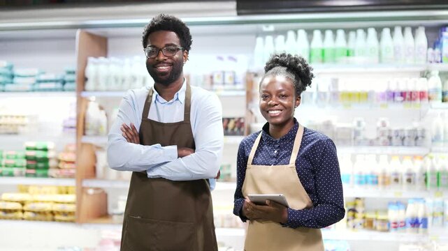 Portrait of two smiling African American family business owners man and woman standing in grocery store behind counter looking at camera. Happy team of food market workers in aprons in dairy departmen