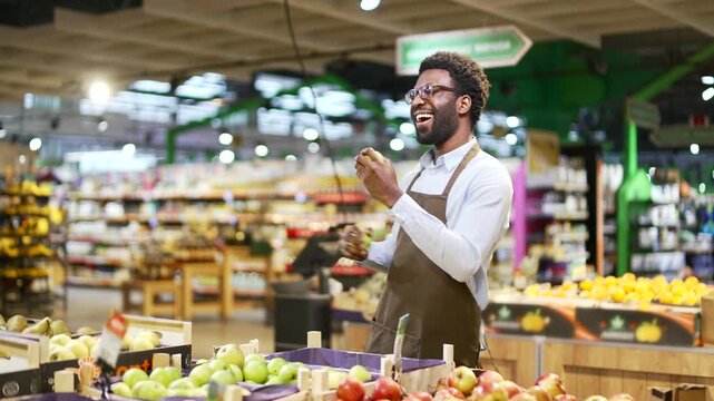 Happy african american male greengrocer dancing at workplace near counter with fruits and vegetables. Salesman in grocery store or food market is having fun. Dance of positive cheerful worker in apron