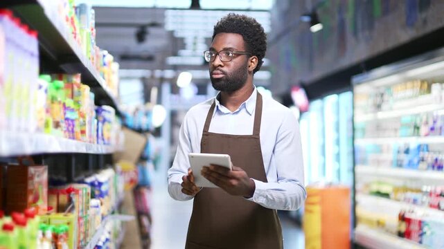 African american male supermarket worker checking availability and condition of products using digital tablet standing in store market. Manager looking at shelves with goods write list inventory