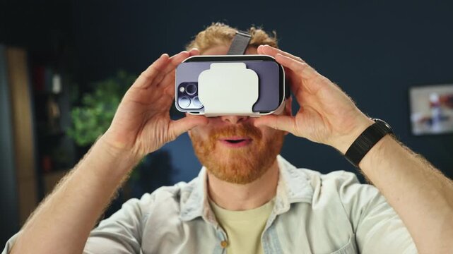 A man is smiling and interacting with a virtual reality headset while sitting indoors. He claps his hands and seems engaged in the experience. The setting appears casual and relaxed.