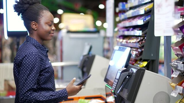 African american female customer using self-service cashier checkout in a supermarket. Shopper scanning produce items using at store self serve cash register pos terminal. Woman banking pay online