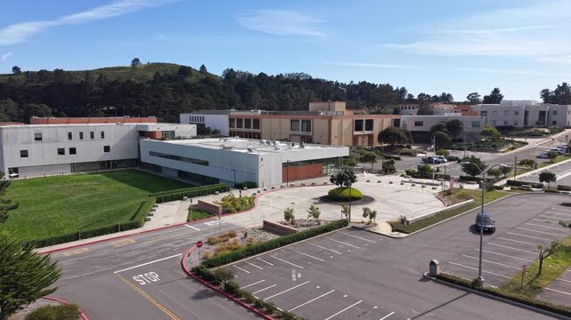 Sunny afternoon aerial view of the Skyline College campus showing modern buildings, a landscaped roundabout, an empty parking lot, and the lush green hills of San Bruno in the background