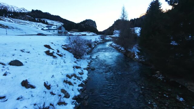 Julierpass, Switzerland, FPV drone fly-forward following a winding river through a snowy alpine valley at dusk, passing roadside slopes and revealing a serene winter mountain landscape