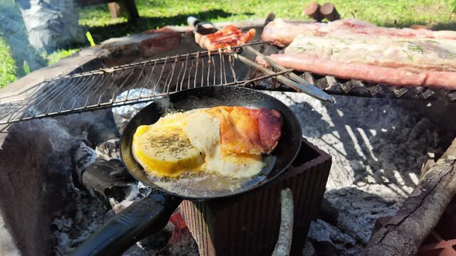 Provoleta cheese melts in a skillet over a rustic outdoor grill. Perfect for culinary content or food culture documentaries showcasing traditional Argentine asado.