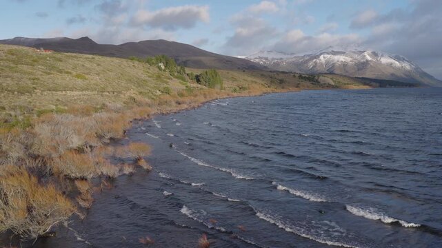 Gentle waves lap at the rocky shoreline of Huechulafquen Lake, framed by snow-capped mountains and rugged terrain. Ideal for travel content or nature documentaries.