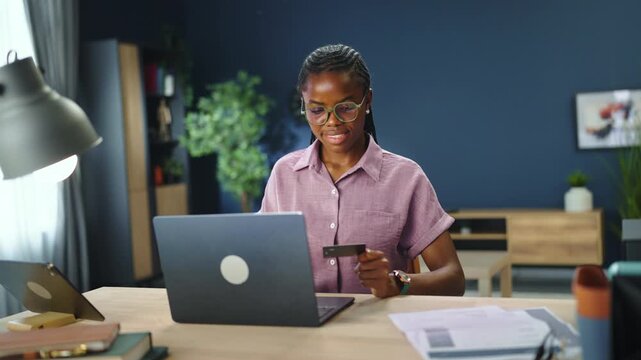 A person is focused on a laptop while holding a phone. The setting is a modern office with plants and a lamp near the workspace. Time appears to be daytime.