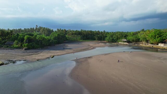 Aerial drone footage of Kelecung black sand beach in Bali, Indonesia, with a wide river coming out and separating the beach into two, green lush vegetations and coconut trees, a very small temple