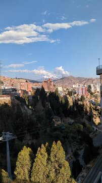 Scenic panoramic view of La Paz city center with green trees, Mi Teleferico infrastructure towers and residential houses on hills under blue cloudy sky with Illimani mountain background, Bolivia