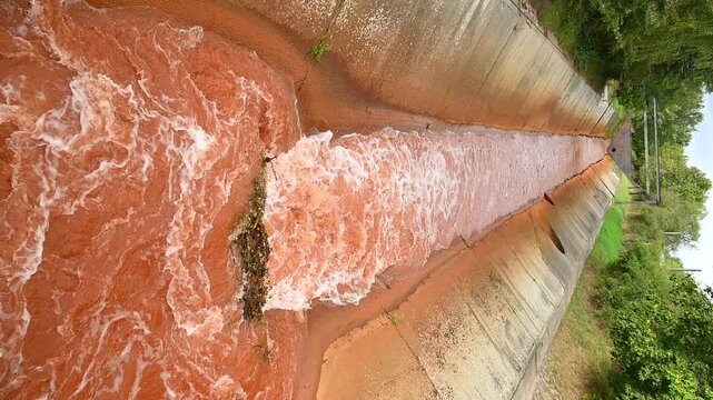 Rust-colored industrial wastewater flowing through a concrete drainage canal near vegetation, showing iron oxide runoff and pollution from iron and steel manufacturing.