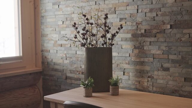 Modern vase with dry branches sitting on a wooden table against a stone wall in a cozy room