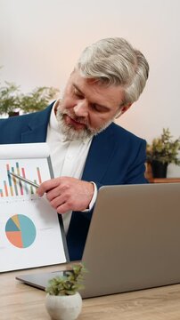 Middle-aged businessman at home office presents graph via laptop meeting noting decline seeking input on. Freelancer guy at table offers comeback strategy gesturing milestones guiding revenue rebound.