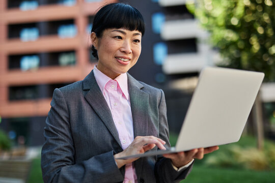 Portrait of a young asian japanese businesswoman woman using a laptop computer and sitting on a bench in a coty park, surrounded by modern corporate office buildings