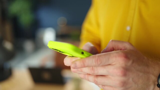 A person is holding a green smartphone and looking at the screen. The setting is a well-lit room with blurred background details. The focus is on the phone and the user&rsquo;s hands.