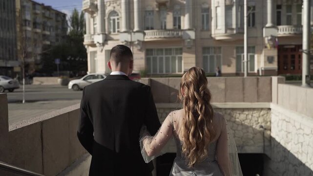 Groom in a black classic suit and bride in silver dress descend the stairs of metro station during the day. Newlyweds are walking through an underground passage in the city.