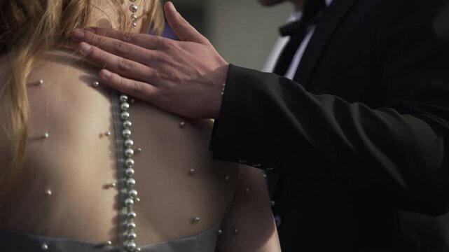 Groom in black suit touches the back of his bride in silver buttoned dress. Close-up of the couple embraces.