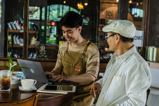 Modern small business digital management young entrepreneur using laptop and tablet planning, finance workflow efficiency technology cafe business operation, learning from senior cafe owner experience