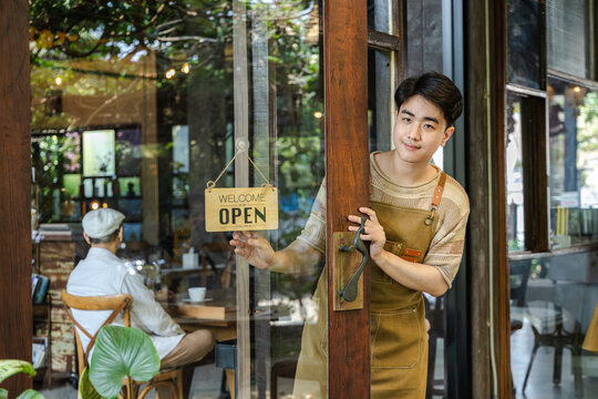 Young barista cafe owner flipping sign to open on storefront door, welcoming customers with confident smile, small business startup energy and positive service mindset in hospitality environment.