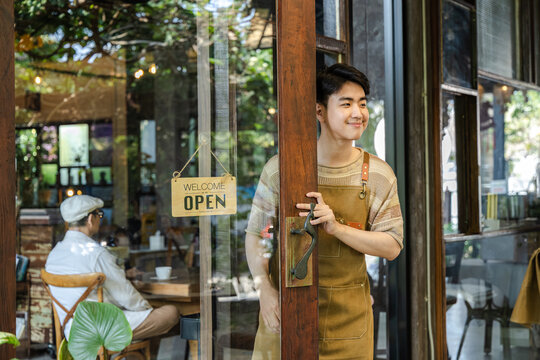 Small business cafe owner barista opening door while holding smartphone welcoming customer into coffee shop showing service interaction and daily business routine, young generation startup business
