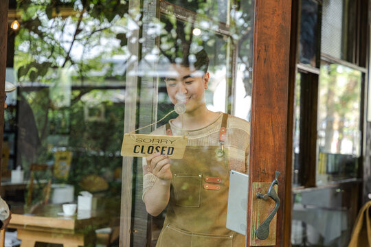 Young small business cafe owner placing closed sign on glass door, young entrepreneur ending daily service routine, reflecting on business operation and customer flow during quiet afternoon moment.