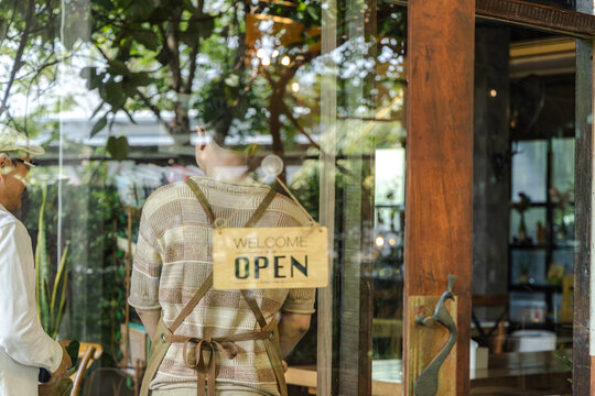 Young small business cafe owner barista open sign welcome gesture standing at entrance inviting customers into coffee shop friendly service concept and positive business presentation, cheerful worker
