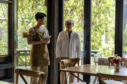Senior cafe small business cafe owner guiding young barista son arranging table seating layout in coffee shop and restaurant, operation planning and mentoring concept in family business environment.