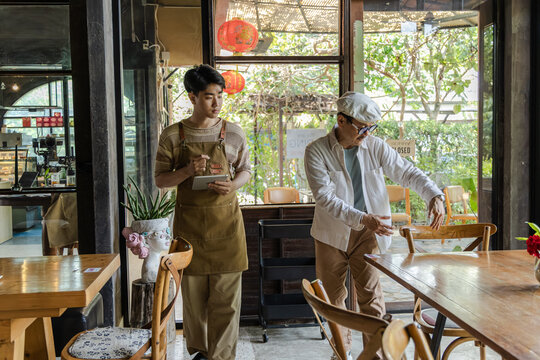 Senior cafe small business cafe owner guiding young barista son arranging table seating layout in coffee shop and restaurant, operation planning and mentoring concept in family business environment.