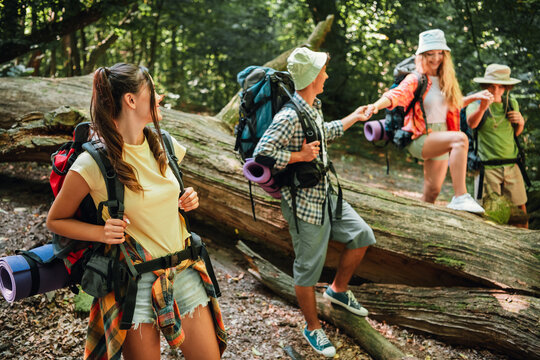 Four friends hiking together in a forest with backpacks and camping gear, helping each other over a fallen log during a summer outdoor adventure