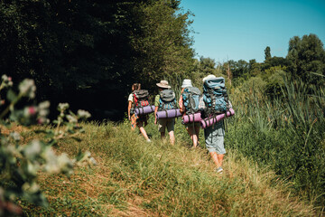 Fototapeta premium Group of hikers backpacking along a grassy trail toward a shaded forest on a sunny summer day