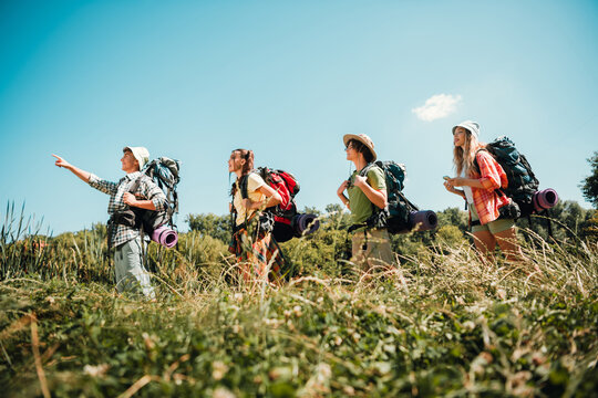 Four friends hiking through a grassy meadow carrying backpacks and camping gear on a sunny summer day exploring nature and outdoor trails