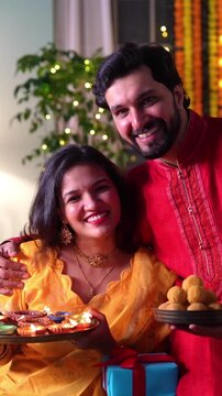 Young Indian couple in stunning yellow and red attires celebrate Diwali in a beautifully decorated home surrounded by diyas, sweets and sharing heartfelt wishes for a happy diwali.