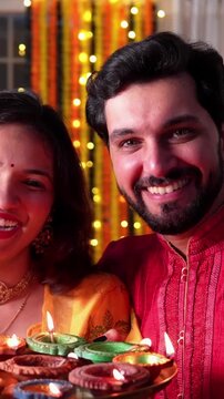 A beautiful happy Indian couple lights Samai and adorns home with colourful painted diyas, enjoying delicious laddoos in a joyous Diwali celebration.