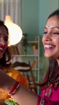 Two beautiful Indian women in traditional bright clothes and jewellery light Samai with oil diyas welcoming Diwali with smiles at home adorned with stunning festive decorations