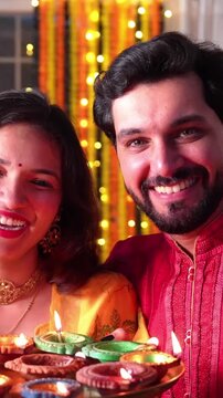 A beautiful Indian couple celebrates Diwali night by lighting Samai and oil diyas, holding laddoos in hand, sharing love, joy and happiness under the glow of festive lights